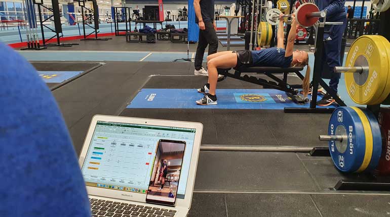 I ett gym, en person tränar bänkpress och i förgrunden sitter en person med en dator och en telefon. I telefonen syns en annan person som tränar på distans.