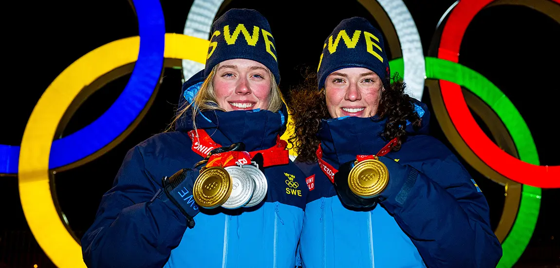 Två leende tjejer i svenska OS-kläder och guldmedaljer runt halsen med OS-ringarna i bakgrunden. 