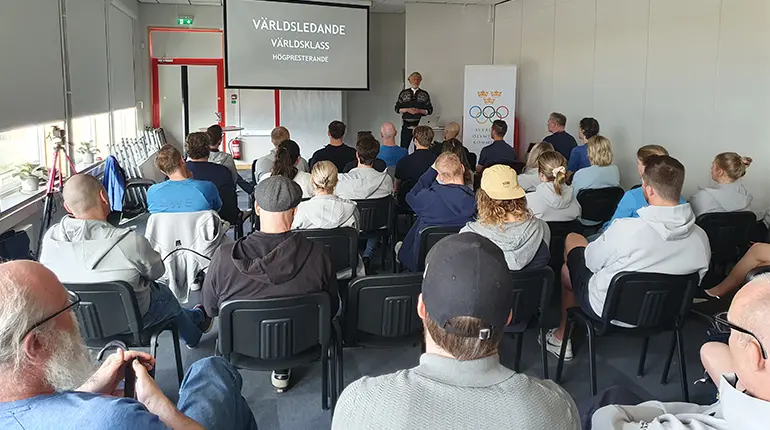 Publik i föreläsningssal. Föreläsaren står framför en presentation med orden Världsledande, Världsklass, Högpresterande. 