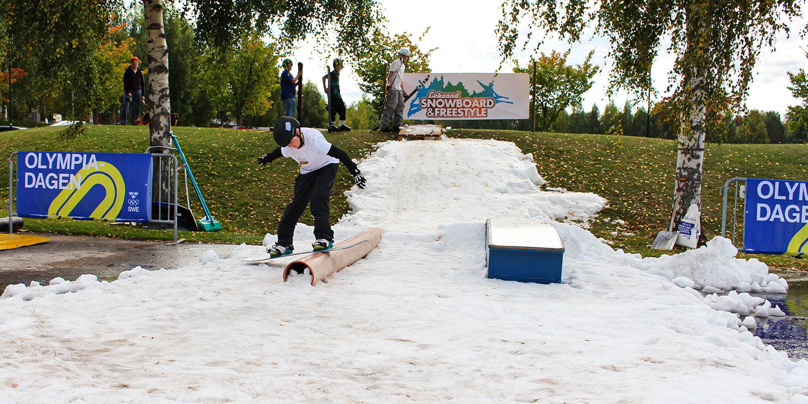 Pojke åker snowboard över ett räcke på snö som ligger på en grön gräsmatta. 
