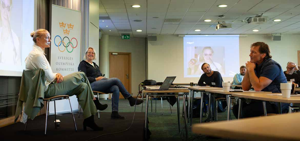 Två personer sitter på ett podium och pratar inför publik.