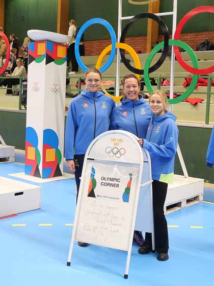 Tre tjejer med blåa och gula tröjor som står framför de olympiska ringarna. 