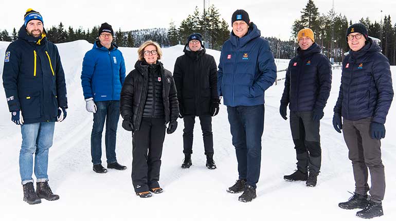 En grupp med herrar och damer i vintermiljö med tjocka jackor och mössor som tittar in i kameran. 
