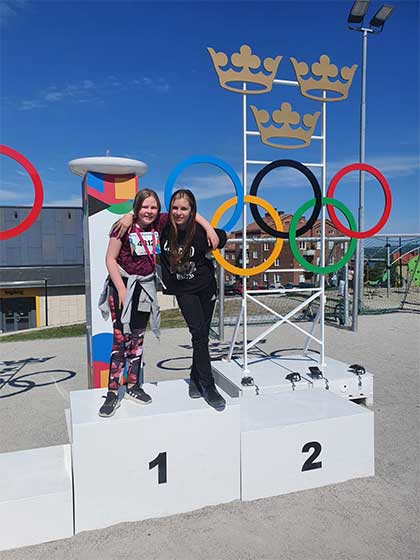 Två flickor som står överst på en stor prispall med de olympiska ringarna i bakgrunden.