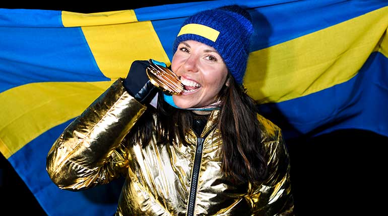 Leende kvinna i guldjacka som biter i en guldmedalj.